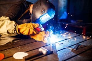 Soldador usando m&aacute;scara de prote&ccedil;&atilde;o, luvas e equipamento de seguran&ccedil;a enquanto realiza soldagem em metal, com fa&iacute;scas vis&iacute;veis na oficina.